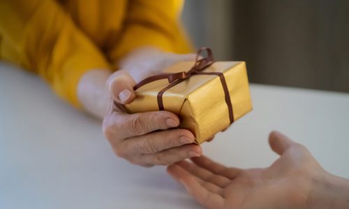 Female hands hold a gift closeup, top view. A young girl gives a nice packed surprise to her mother, grandmother with love, remembers elderly relatives, visits during holidays, takes care of them.
