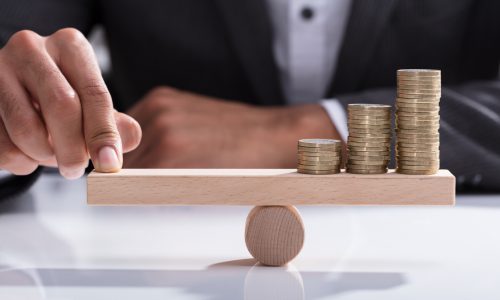 Close-up Of A Businessperson's Hand Balancing Stacked Coins On Wooden Seesaw With Finger Over Desk